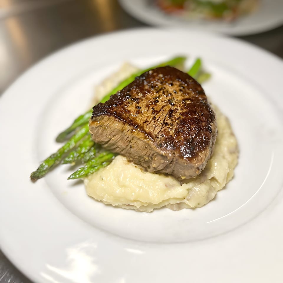 Seared steak with mashed potatoes and green asparagus spears on a white plate.