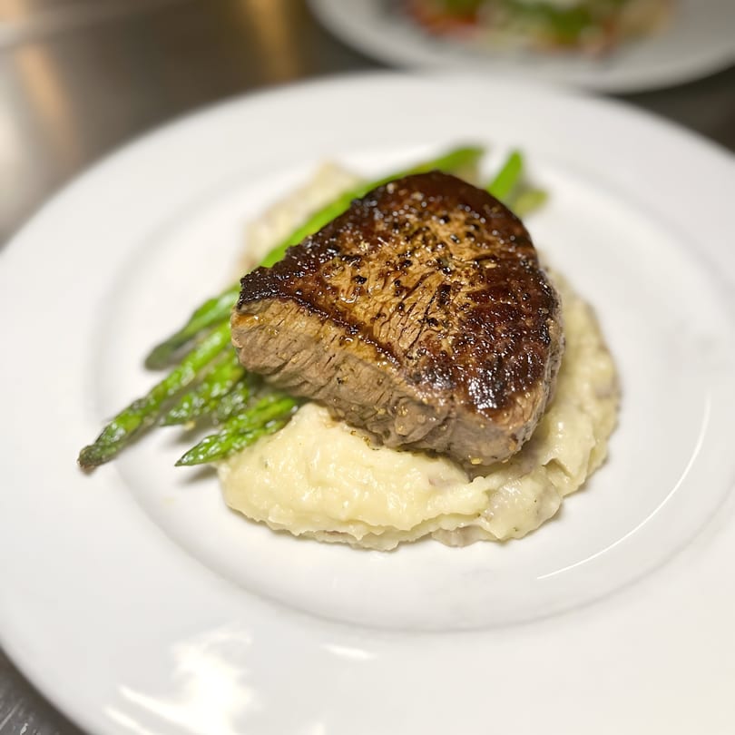Seared steak with mashed potatoes and green asparagus spears on a white plate.