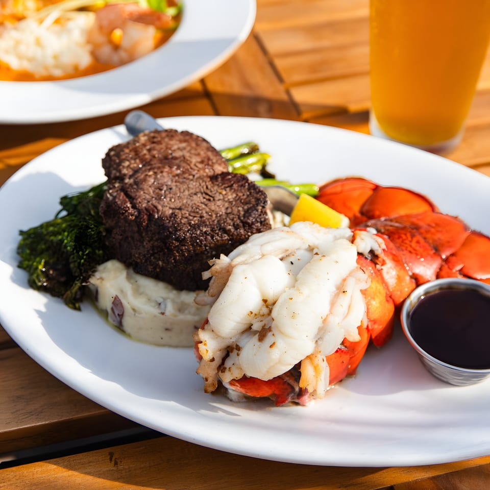Mad Fish surf and turf with steak, lobster tail, mashed potatoes, and greens on a white plate.